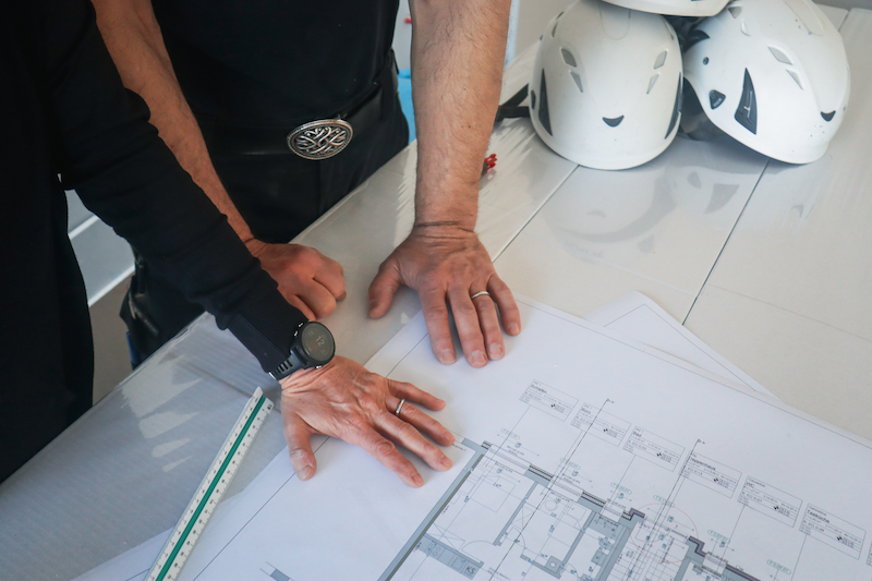 Zwei Personen beugen sich über einen Bauplan auf einem Tisch, daneben liegen drei weiße Schutzhelme und ein Lineal – Symbol für Zusammenarbeit auf der Baustelle.
