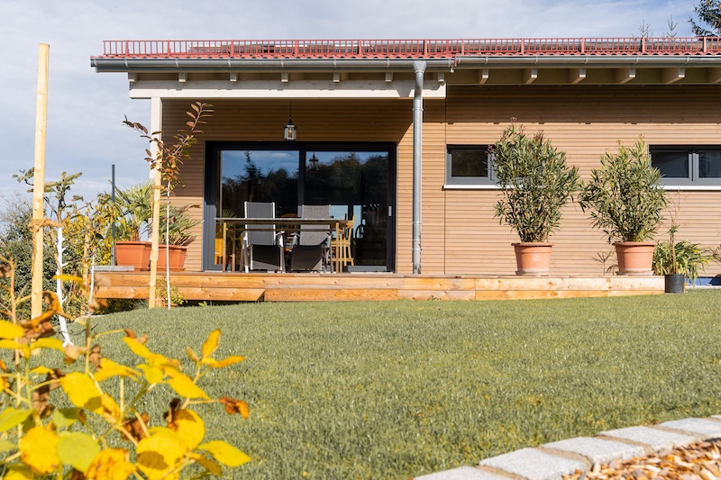 Frontansicht eines modernen Einfamilienhauses mit horizontaler Holzverschalung, bodentiefer Schiebetür zur Terrasse, Topfpflanzen und Sitzgruppe – davor ein gepflegter Garten mit Rasen und jungen Bäumen.