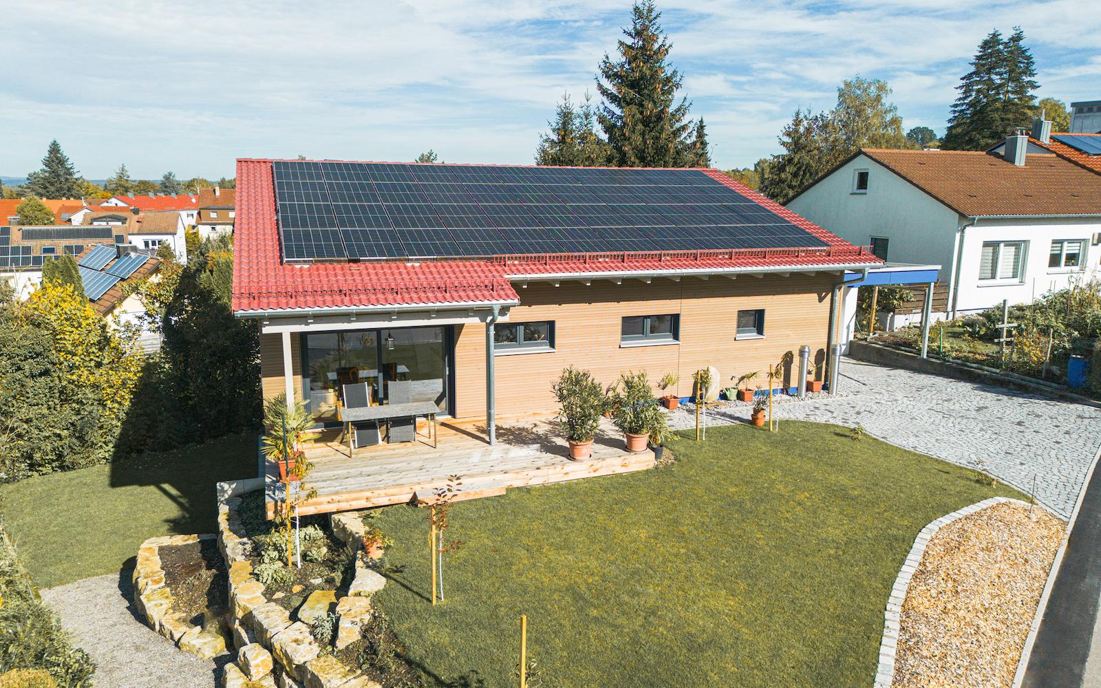 Gesamtansicht eines Einfamilienhauses mit rotem Ziegeldach, Photovoltaikanlage, Holzfassade und großzügiger Terrasse im gepflegten Garten.