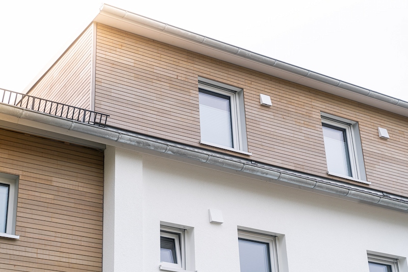 Detailaufnahme eines aufgestockten Gebäudes mit horizontaler Holzverschalung im oberen Geschoss und verputzter weißer Fassade darunter. Zwei Fenster mit Aluminiumrahmen durchbrechen das ruhige Fassadenbild.
