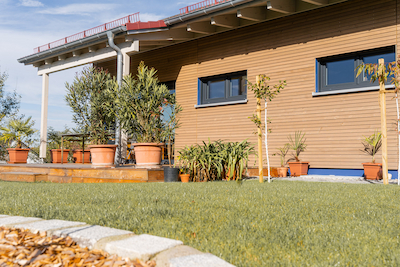 Ansicht einer modern gestalteten Hausfassade mit horizontaler Holzverkleidung, zwei dunkel gerahmten Fenstern und vorgelagerten Pflanzkübeln auf einer Terrasse – davor gepflegter Rasen und junge Bäume.
