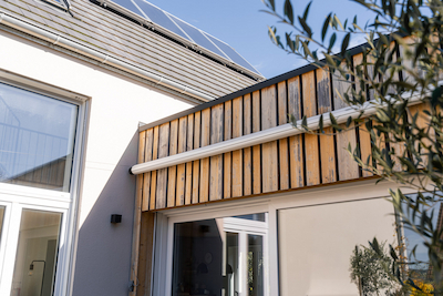 Detailansicht eines modernen Anbaus mit vertikaler Altholzverschalung über bodentiefen Fenstern, daneben glatte Putzfassade und Solarmodule auf dem Dach – nachhaltige Gestaltung mit Materialkontrast.