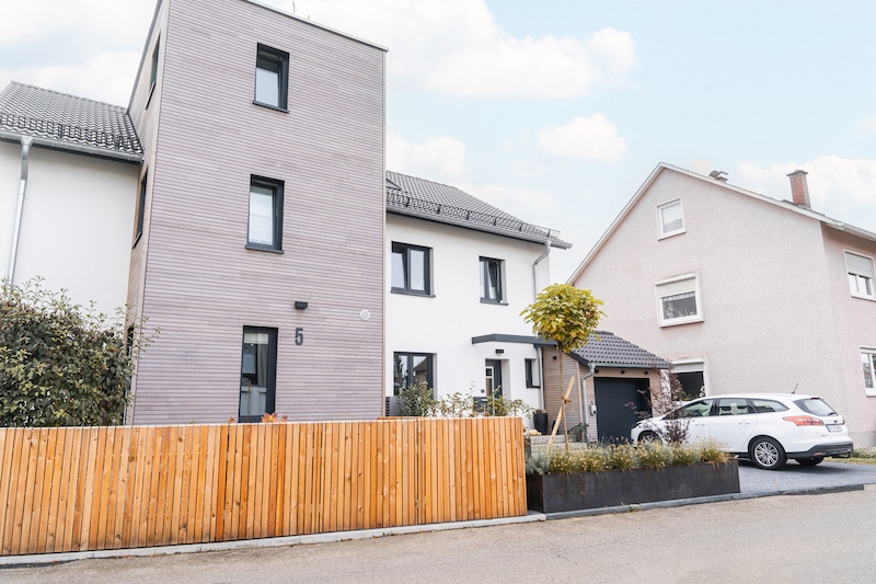 Vorderansicht des Hauses mit senkrechter Holzverkleidung und dunkelgrauen Fenstern, umgeben von gepflegtem Garten.