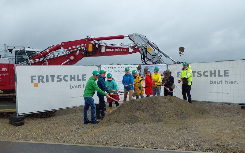Feierlicher Spatenstich mit Projektbeteiligten vor einem Bagger und Baustellenbanner auf dem Baugelände.
