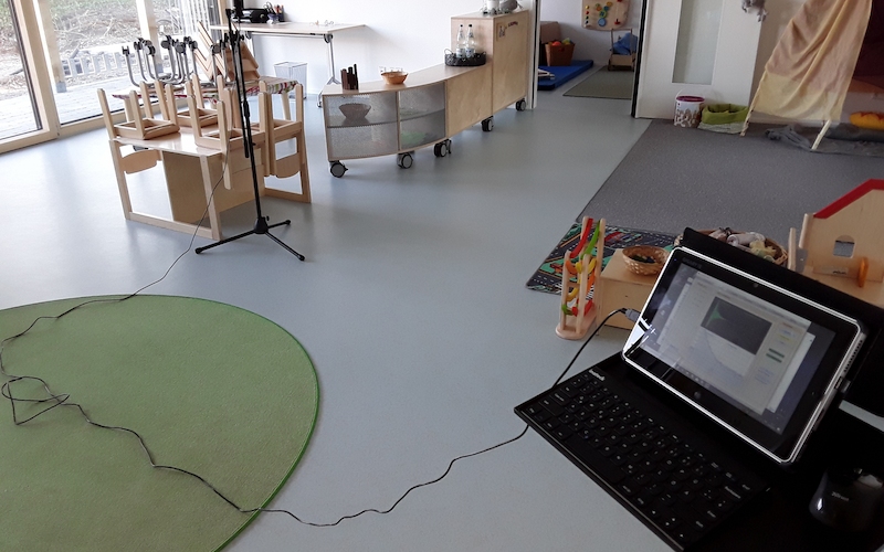 Heller Gruppenraum mit kindgerechten Möbeln, Teppich und Tablet-Setup für akustische Messung.