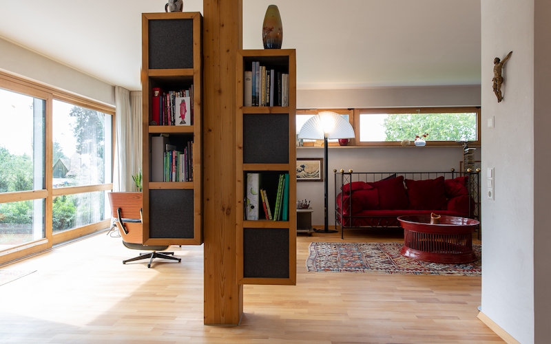 Wohnraum mit heller Fensterfront, freistehendem Holzregal und gemütlicher Sitzecke mit rotem Sofa und Teppich.