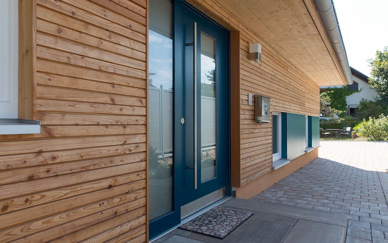 Moderner Hauseingang mit Holzfassade, barrierefrei zugänglicher Haustür und gepflastertem Vorplatz.
