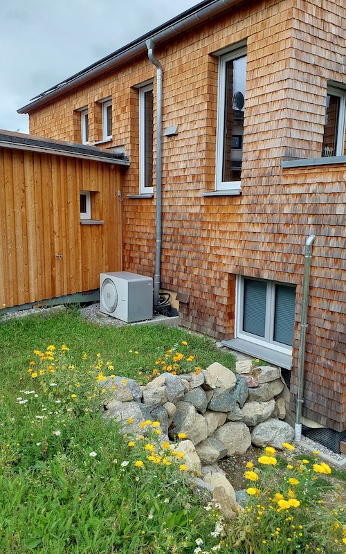 Wohnhaus mit Fassade aus Holzschindeln, Wärmepumpe im Garten und bepflanztem Natursteinbeet.