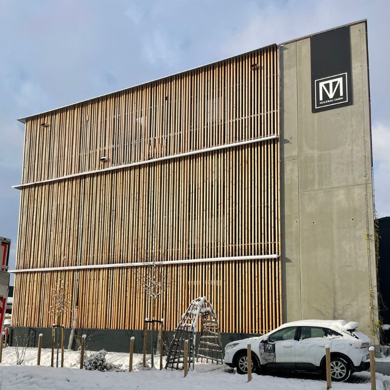 Moderne Gebäudefassade mit vertikaler Holzverschalung und Sichtbeton, winterliche Schneelandschaft im Vordergrund, geparktes Auto mit Firmenlogo – Beispiel für nachhaltige, zeitgemäße Architektur.